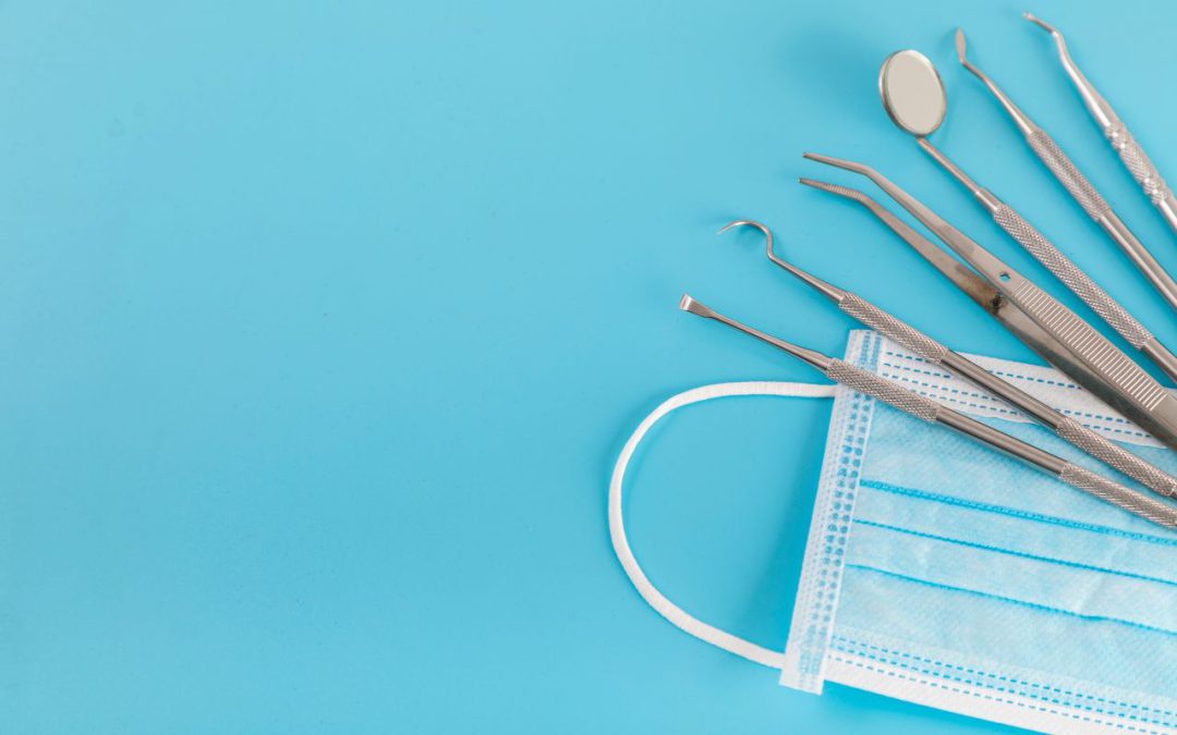 Strumenti odontoiatrici in acciaio e mascherina chirurgica su sfondo azzurro, simbolo di igiene e professionalità in ambito dentistico.